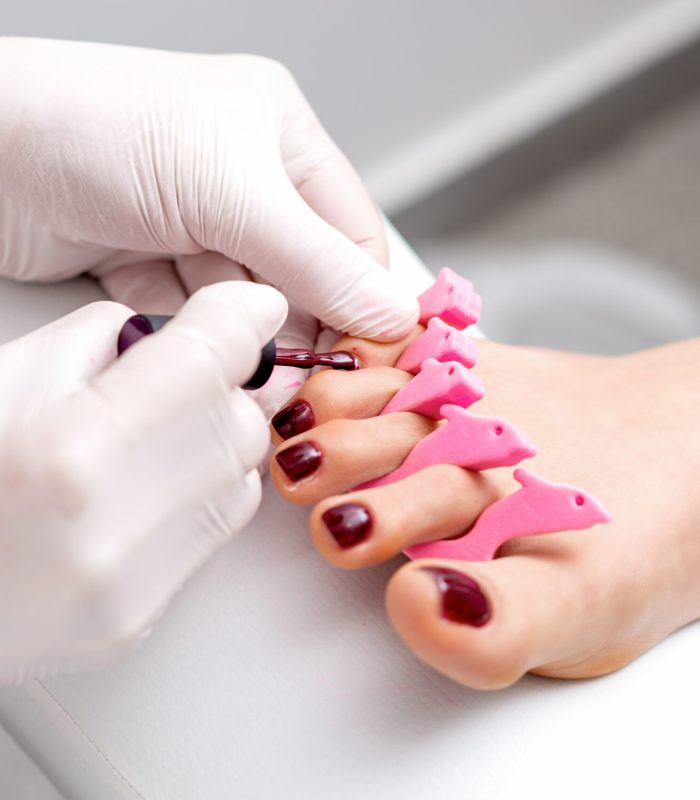 Manicure master is painting on female toenails with maroon nail polish by brush wearing white gloves